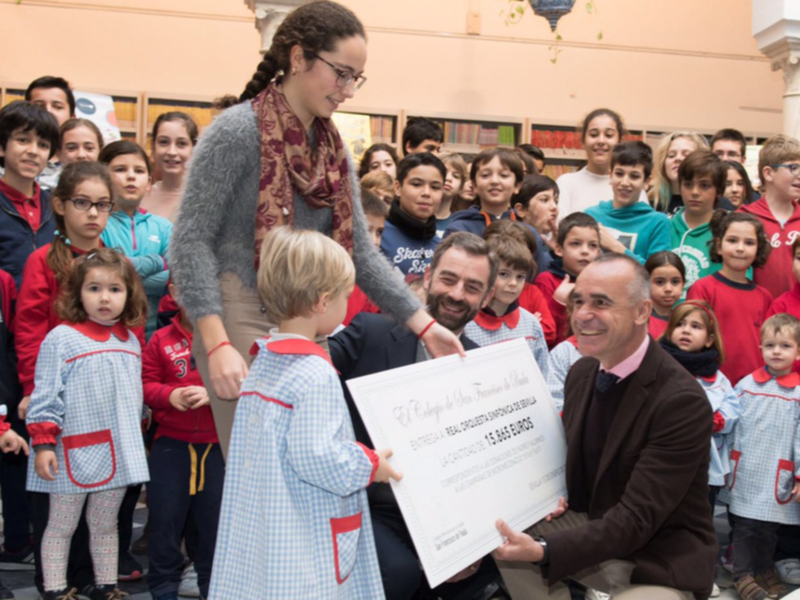 Entrega de un cheque a un niño en presencia de un grupo de niños y adultos en un ambiente escolar.