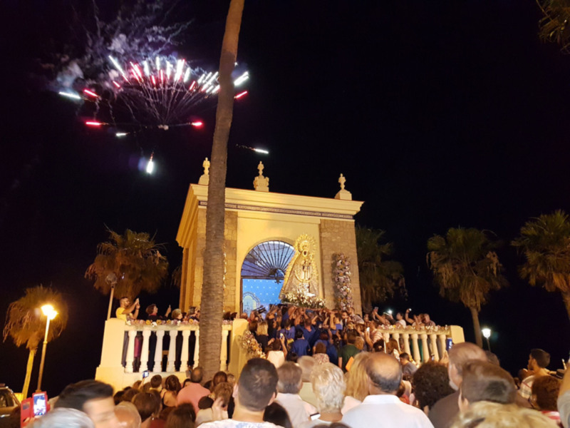 Fuegos artificiales sobre una estatua religiosa en un evento nocturno. La gente disfruta de la celebración, con palmeras y luces en el fondo.