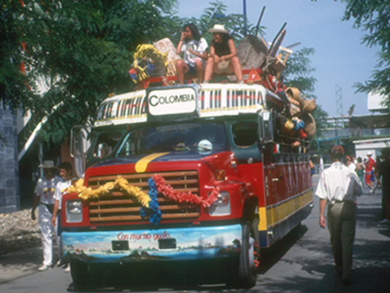Un bus colorido con la bandera de Colombia en el techo, lleno de personas y decorado con animales. La imagen muestra un ambiente festivo en una calle con árboles y edificios al fondo.