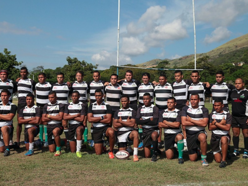 Equipo de rugby posando en el campo con un balón de rugby en primer plano, con montañas y cielo azul de fondo.