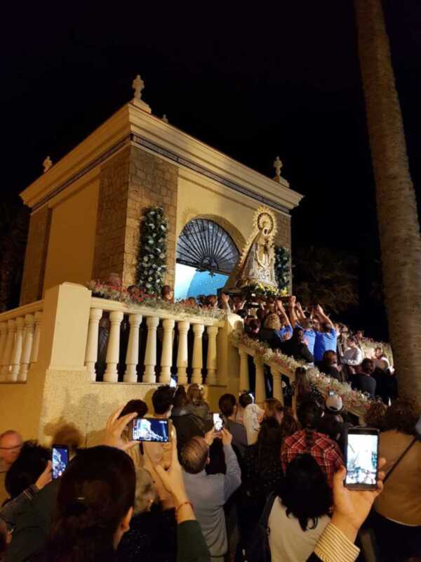 Fiesta religiosa nocturna con procesión y multitud en honor a la Virgen, iluminada por faroles y cámaras de fotos al frente.