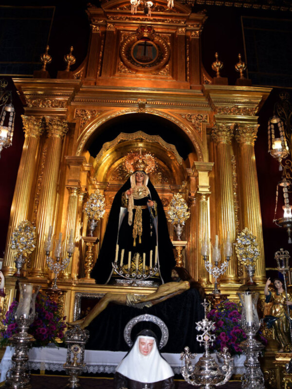 Un altar dorado elaboradamente decorado, con una figura central vestida de negro y rodeada por adornos florales. En el fondo, se observan detalles dorados y una escultura más pequeña a la derecha. La atmósfera es reverente, típica de un espacio religioso.