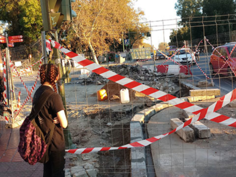 Un hombre y una mujer observan con atención un área de construcción bloqueada por barreras rojas y blancas, mientras una camioneta de trabajo circula en el fondo.