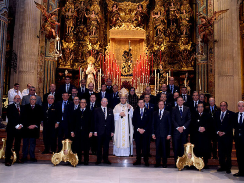 Una imagen de una iglesia con un altar dorado y estatuas, donde un grupo de hombres en trajes formales se encuentra frente a la cámara. El altar está iluminado con velas y hay un ángel en el techo.