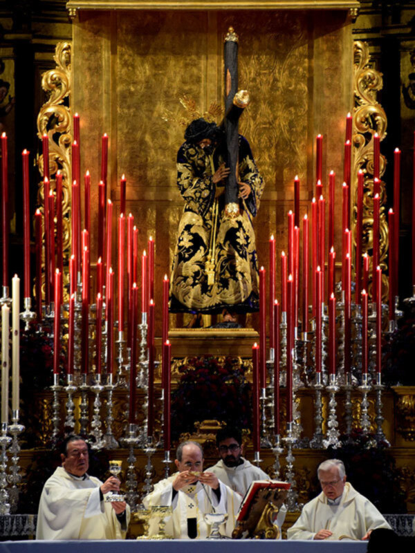 Eucaristía con sacerdotes en el altar y estandarte de Jesús Nazareno en el fondo, rodeado de velas rojas.