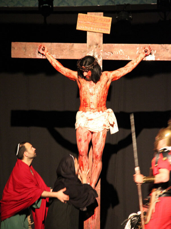 Una escena de la crucifixión con un personaje en la cruz, rodeado por otros actores vestidos de rojo y negro. La escena se realiza en un escenario con una pantalla negra al fondo, iluminada por luces de teatro. La imagen transmite una sensación dramática y religiosa.