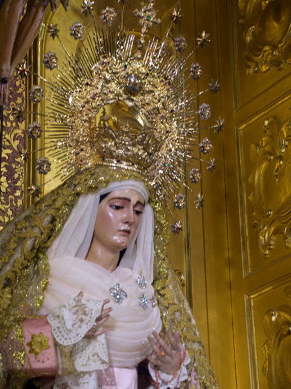 Estatua de la Virgen María con un vestido blanco y una corona dorada, rodeada por detalles dorados en el fondo.
