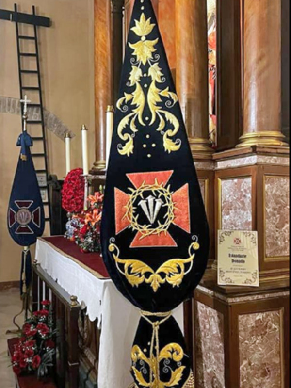 Imagen de una bandera ceremonial colgada en un altar, con detalles dorados y rojos sobre fondo negro. La decoración es elaborada y simbólica, posiblemente perteneciente a una institución religiosa o militar.