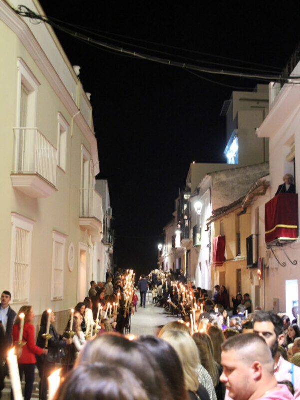 Fotografía nocturna de una procesión religiosa con personas sosteniendo velas en una calle estrecha rodeada de edificios blancos y amarillos, con un cielo oscuro y algunas luces de farola en el fondo.