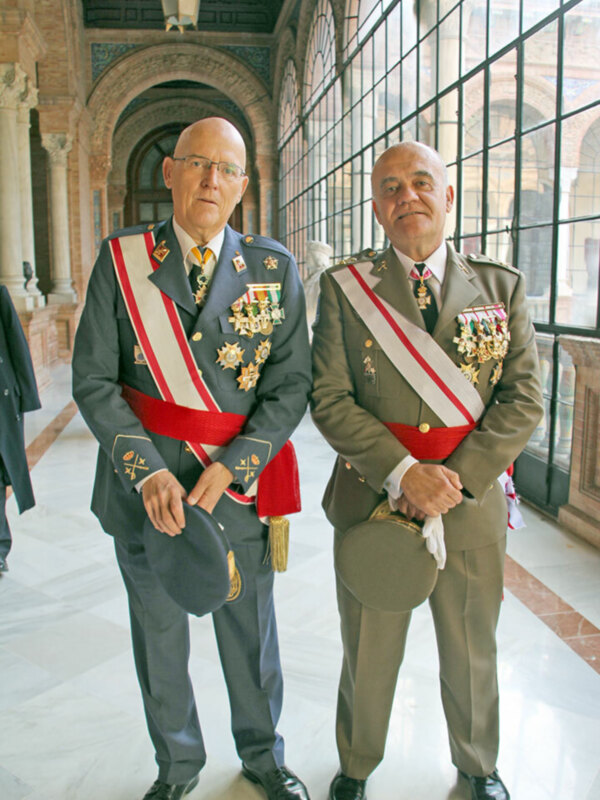 Dos hombres en uniforme militar posan frente a una ventana grande con persianas. El hombre de la izquierda lleva un uniforme azul con insignias y una corbata roja, mientras que el hombre de la derecha lleva un uniforme verde con insignias y una corbata roja. Ambos tienen medallas en sus uniformes. El fondo muestra un interior con arcos y columnas de piedra.