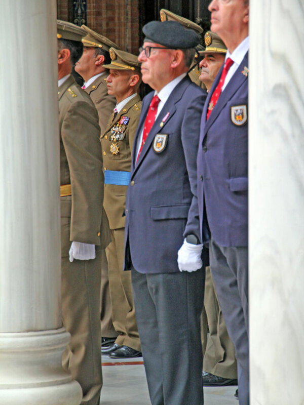 Un grupo de hombres vestidos con uniformes militares y civiles, posiblemente en una ceremonia o evento formal. La imagen muestra a los hombres de pie, algunos con gorras militares y otros con corbatas rojas. La escena parece ser al aire libre, posiblemente frente a una estructura histórica o monumental.