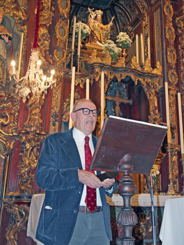 Un hombre en traje y corbata, con gafas, está frente a un altar decorado con estatuas y velas.