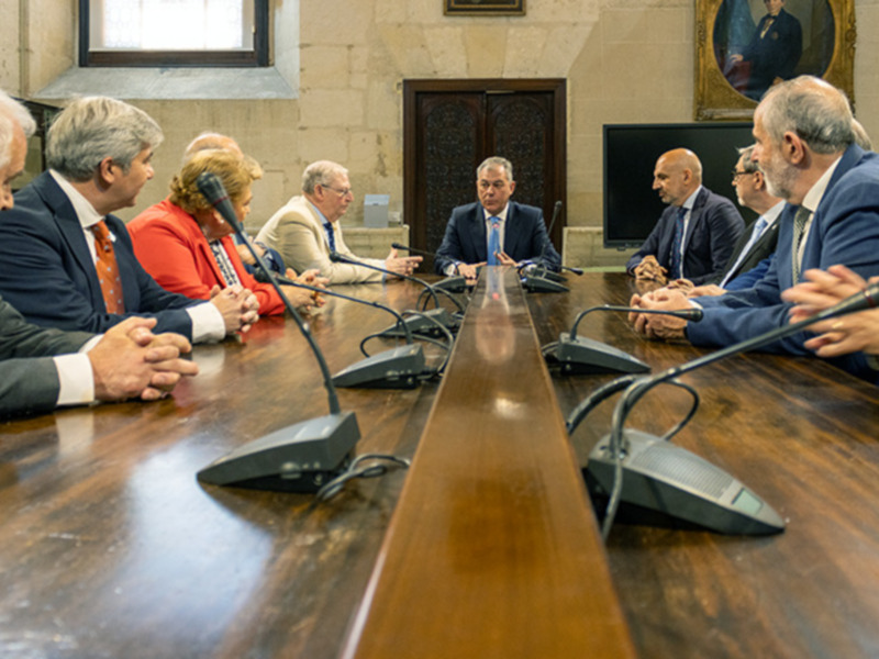 Una reunión formal en un salón de conferencias con personas sentadas alrededor de una mesa de madera, sosteniendo discusiones. La sala tiene un ambiente formal con paredes de ladrillo y una pintura en el fondo. La iluminación es suave, creando un ambiente serio y concentrado para la conversación.