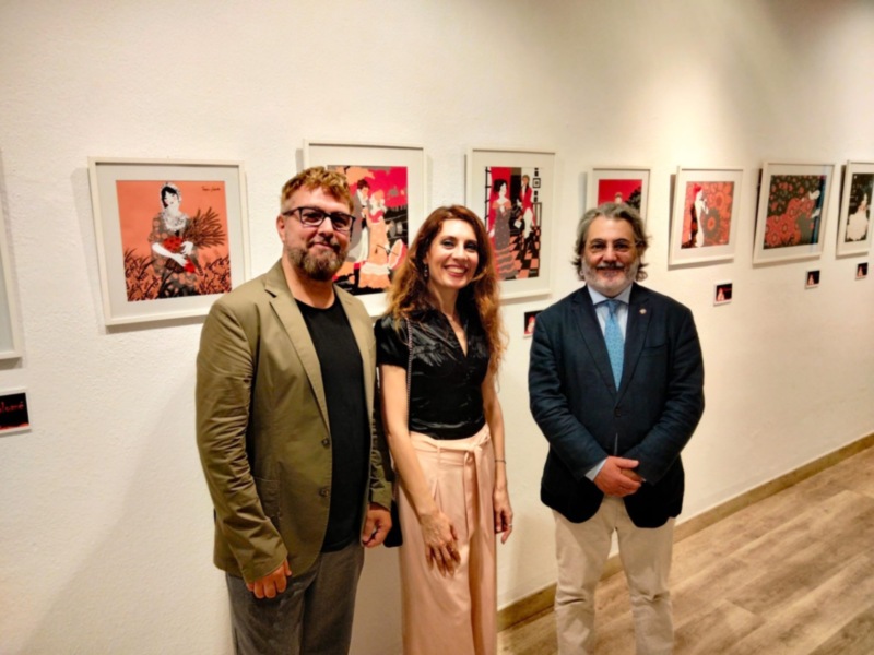 Se observan tres personas posando frente a una pared con varias obras de arte. La escena parece ser parte de una exposición o evento artístico.