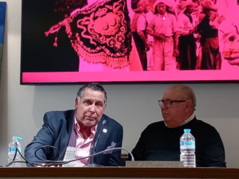 Una reunión formal con tres personas sentadas en una mesa, frente a un televisor que muestra imágenes de una procesión religiosa. La mesa tiene botellas de agua y microfonos, indicando una conferencia o presentación.