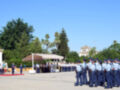 Fotografía de una ceremonia militar en un parque con palmeras y árboles, donde soldados en uniforme se alinean en formación. El cielo es azul claro y hay una estructura con una cruz en el fondo. La ceremonia parece ser formal y ceremonial.