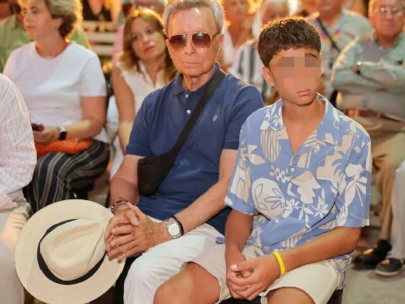 Un hombre adulto y un niño sentados en una audiencia, con el hombre usando gafas de sol y la mujer de fondo con un vestido blanco.