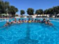 Un grupo de jóvenes jugando voleibol en una piscina con un balón amarillo. El cielo es azul claro y hay árboles alrededor de la piscina.