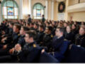 Un grupo de personas en uniforme militar sentadas en filas. La imagen muestra una sala con vitrales y un escudo decorativo en el fondo.