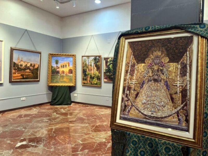 Una sala de exposición con varias pinturas, una destacada en el centro. La imagen muestra una figura vestida de manera elaborada con detalles dorados y azules, posiblemente un personaje histórico o mitológico. La pared al fondo muestra una vista de un edificio colonial con palmeras, sugeriendo un tema histórico o cultural. La iluminación es suave y el suelo tiene un patrón de cuarzo rojo.