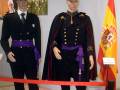 Dos modelos de uniformes militares, uno con un sombrero y otro con una capa, frente a una bandera española.