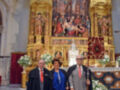 En el fondo, un altar dorado con esculturas y una pintura central que muestra la crucifixión. En el frente, tres personas posan frente al altar, con un hombre en traje y sombrero, una mujer con abrigo azul y bastón, y otro hombre en traje gris. A la izquierda del altar, flores blancas adornan el suelo.