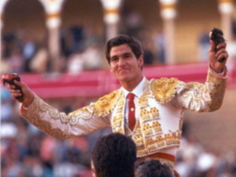 Un torero en traje tradicional, con un fondo de arcos dorados y espectadores en la arena.