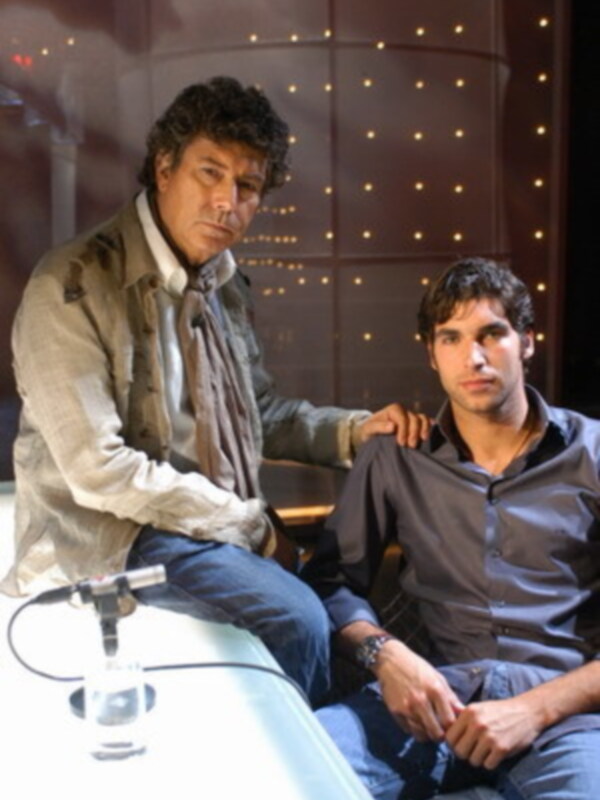 Dos hombres en un entorno de luces doradas, uno está sentado y el otro está ayudándolo con un objeto.