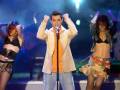 Tres personas en un escenario, dos mujeres con trajes de baile y un hombre con una camisa blanca y pantalones azules, todos con gestos de celebración.