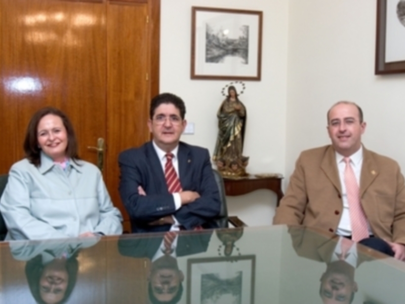 Tres personas sentadas en una oficina, con un retrato de la Virgen María en el fondo y una mesa de cristal en el centro.