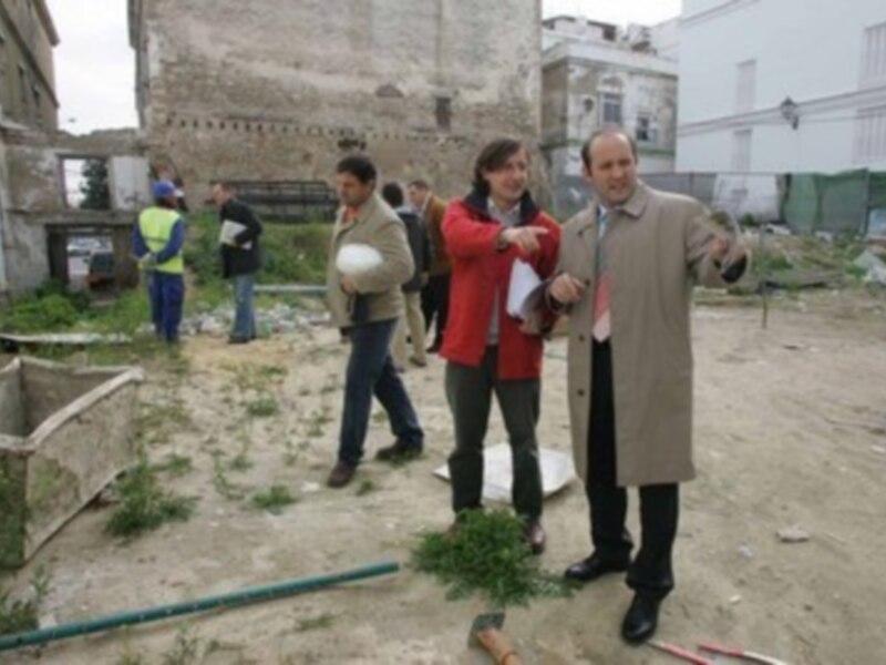 Dos hombres en una zona de construcción, uno de ellos sostiene un pañuelo blanco. A su lado, hay un edificio de ladrillo y una estructura de hormigón. El suelo es despejado, con algunas plantas y una tubería verde en el fondo.