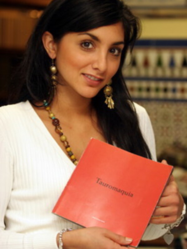 Una mujer con cabello largo y negro, usando un collar de cuentas y pendientes dorados, sostiene una carpeta roja con el título "Tauromagia".
