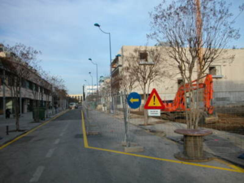Un camino vacío con señales de tráfico, incluyendo un letrero azul con flecha hacia el lado izquierdo y uno amarillo con una figura de tráfico. La imagen muestra un entorno urbano con edificios y árboles sin hojas.