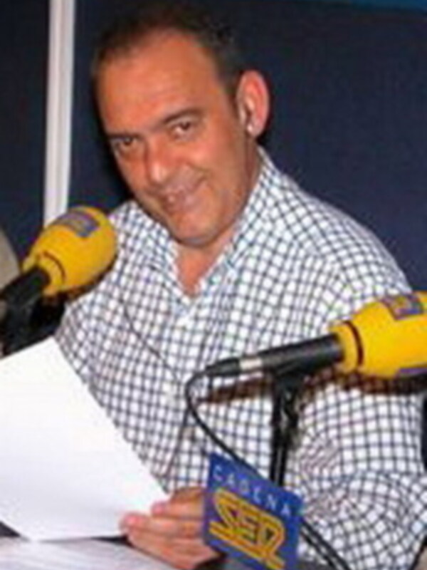 Un hombre sonríe mientras habla en un micrófono de radio, con una pancarta que dice "Radio Sevilla" y un fondo azul.