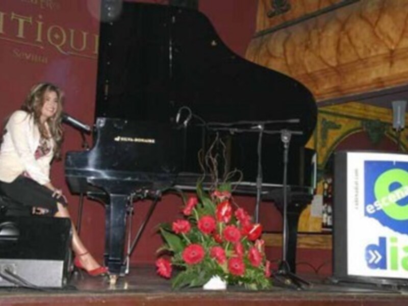 Una mujer está frente a un piano negro en una sala de conciertos, con flores rojas y un cartel de "Esenoti" en el fondo.