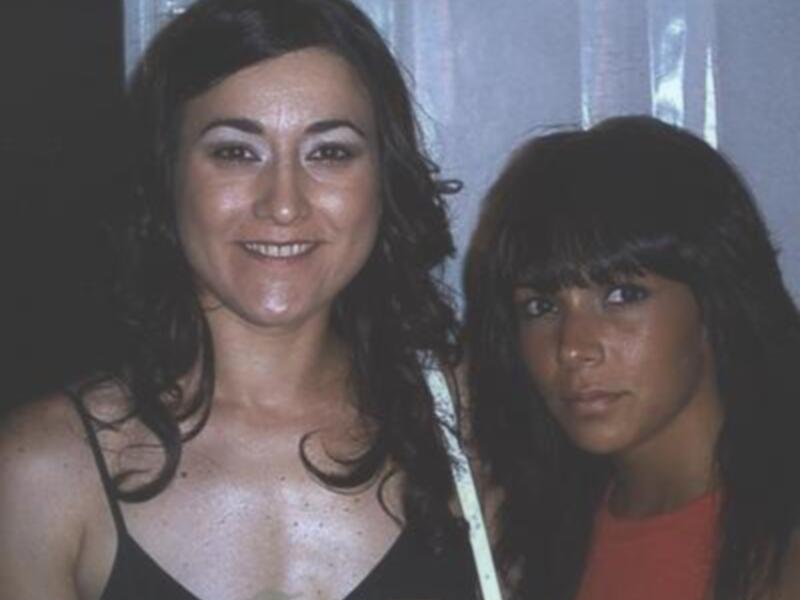 Dos mujeres con cabello largo y oscuro, una en un vestido negro y la otra en un rojo brillante. Ambas tienen expresiones serias y miran directamente a la cámara. El fondo es oscuro, lo que destaca sus rostros y vestidos.