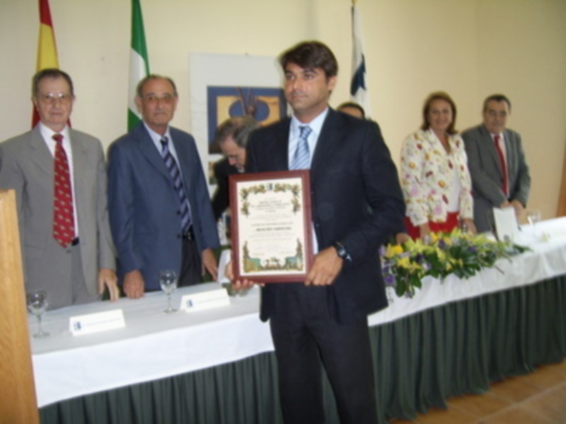 Un hombre sostiene un diploma mientras otros tres hombres y una mujer observan. La imagen se centra en la celebración de un premio o reconocimiento, con una mesa decorada y banderas al fondo.