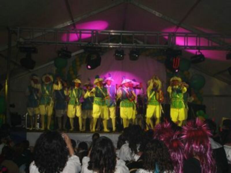 Grupos de música en un escenario iluminado con luces rojas y amarillas, vestidos de colores vibrantes.