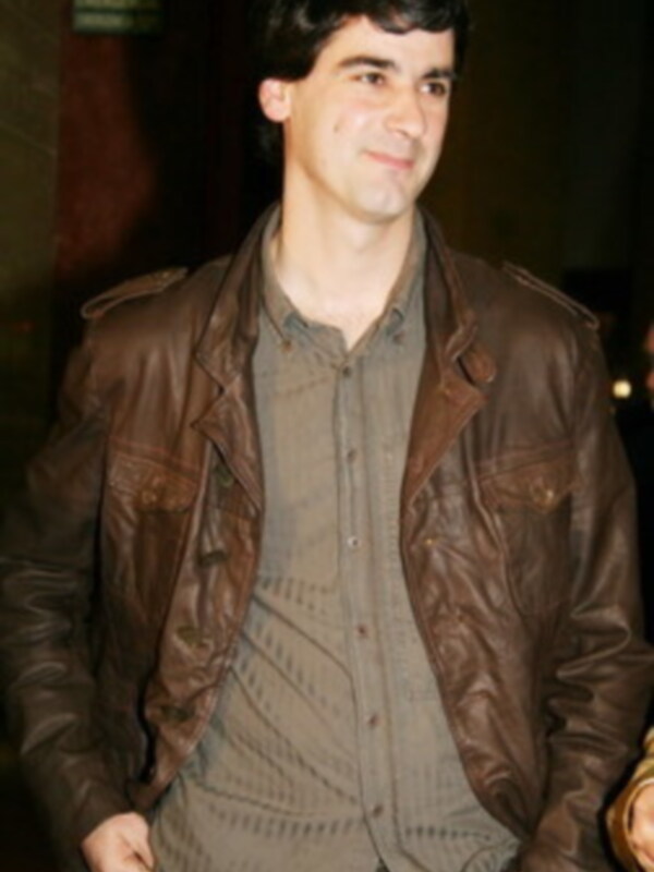 Un hombre con cabello oscuro y barba, lleva una chaqueta de cuero marrón sobre un jersey gris. La imagen parece ser nocturna, posiblemente en una sala de cine o teatro.