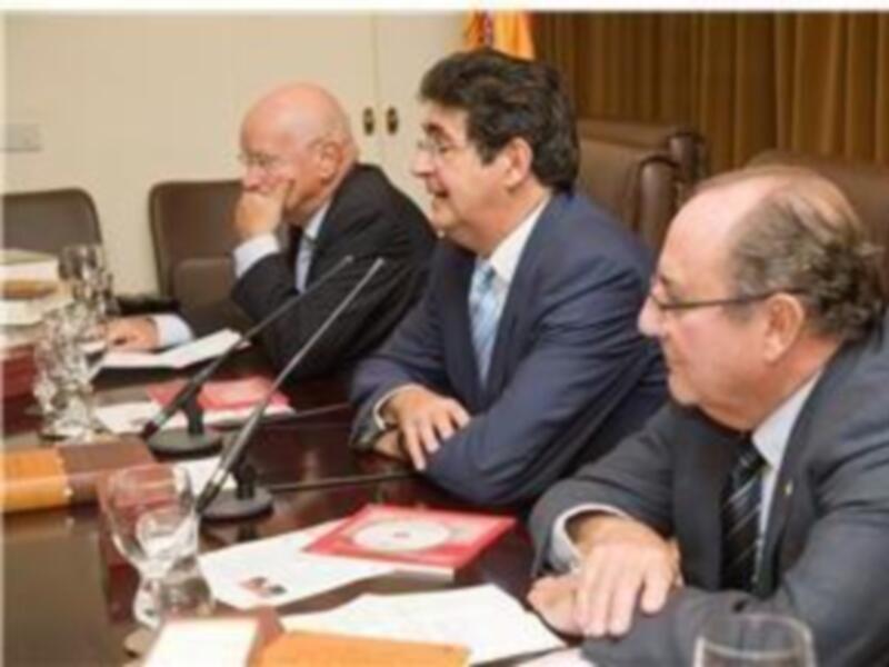 Tres hombres en trajes de negocios sentados en una mesa con micrófonos, probablemente en una reunión o conferencia.