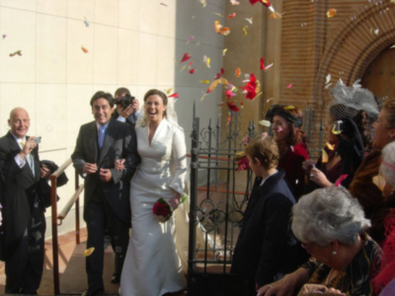 Una boda en un edificio blanco con confeti caído. La novia sonríe mientras el novio la abraza, rodeados por amigos y familiares.