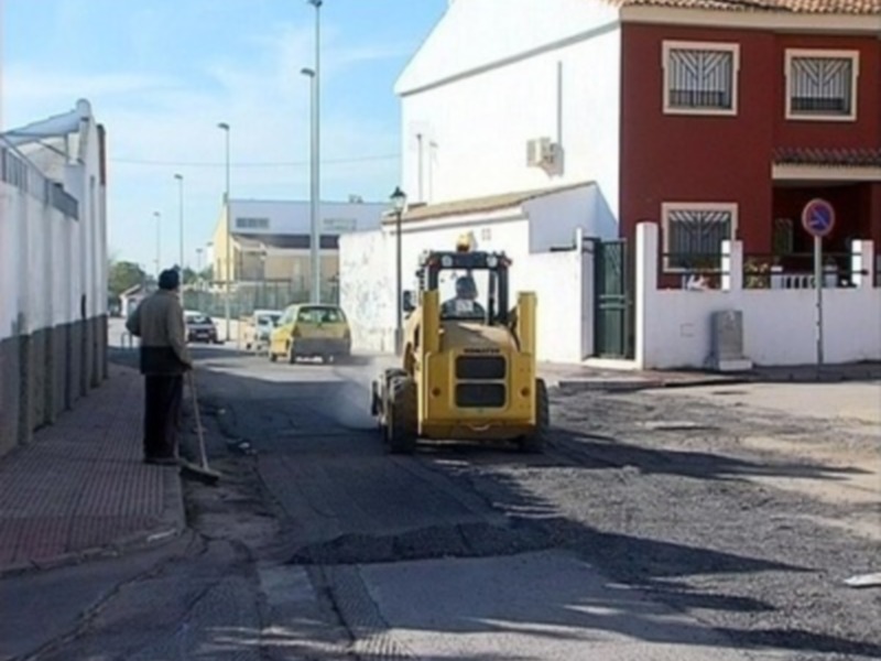 Un trabajador en un camión compactador de asfalto en una calle de una ciudad, con edificios y una señal de tráfico en el fondo.