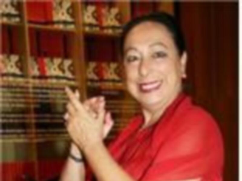 Una mujer con vestido rojo y joyería, sentada y apoyada en un sofá con cojines rojos y negros.