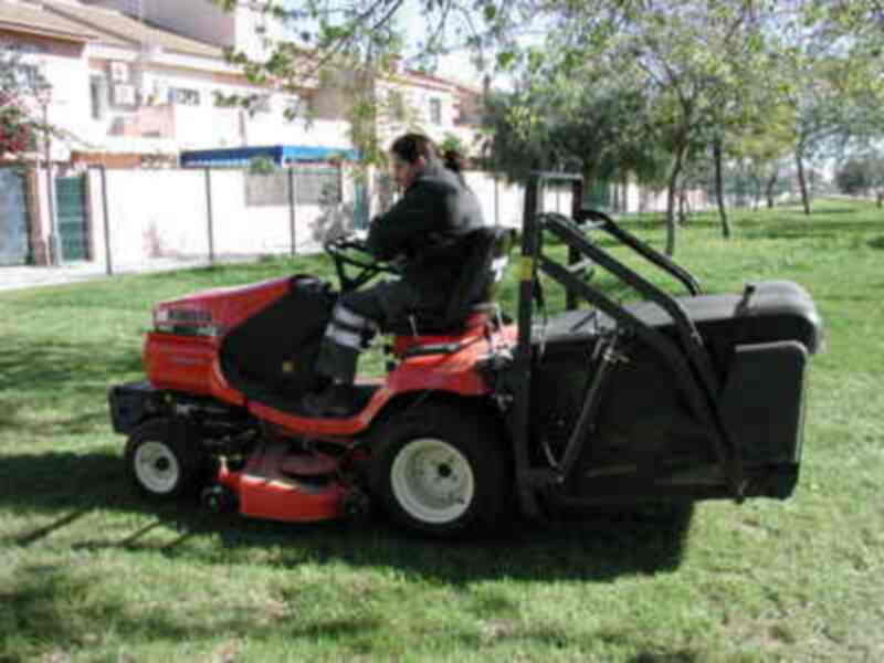 Una máquina de podar en un parque, con un hombre en la conducción. El parque tiene césped verde y árboles al fondo. La máquina es roja y tiene un asiento para el operador. La imagen muestra una escena de mantenimiento del parque.