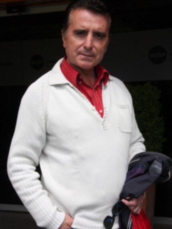Un hombre con una chaqueta blanca y un camisa roja, posando frente a una pared negra con letras blancas.