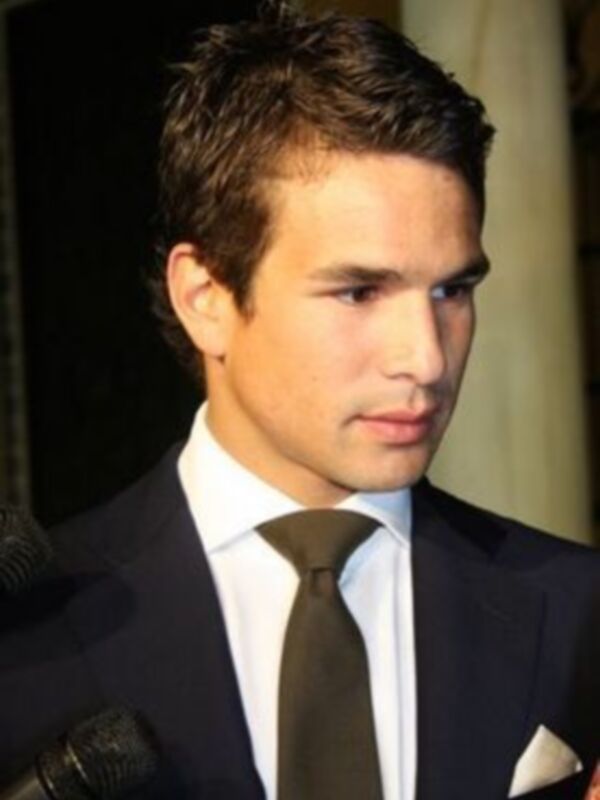 Un hombre con cabello oscuro y peinado en moño, vestido con traje y corbata negra, hablando en un micrófono.