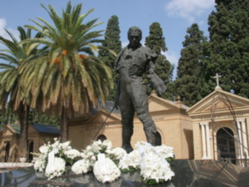 Escultura en piedra de un hombre con una corona, rodeada por palmeras y flores blancas. En el fondo, un edificio con una cruz en la parte superior.