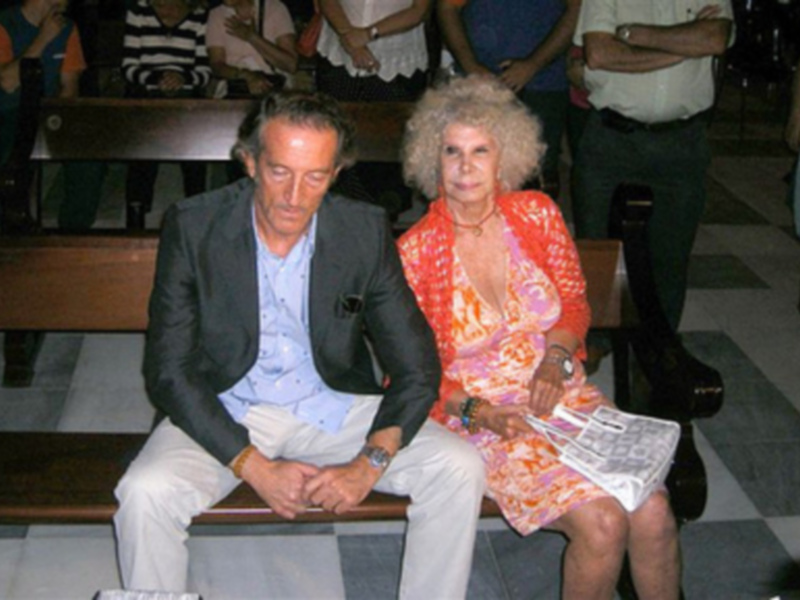 Persona en traje sentada junto a otra en vestido colorido, ambos en un entorno de audiencia, con periódico en las manos.
