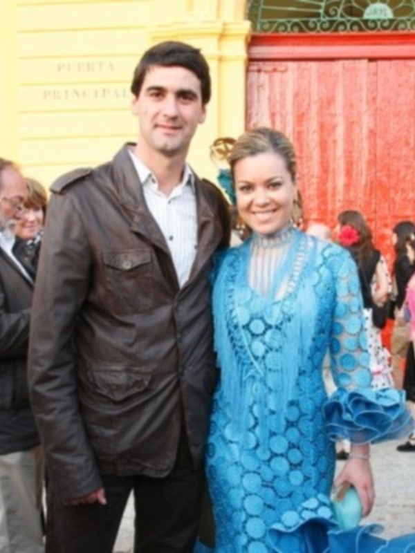 Una pareja se posa frente a la Puerta Principal de un edificio con detalles ornamentales, vestida con un traje tradicional de flamenca azul y el hombre con una chaqueta de cuero marrón.