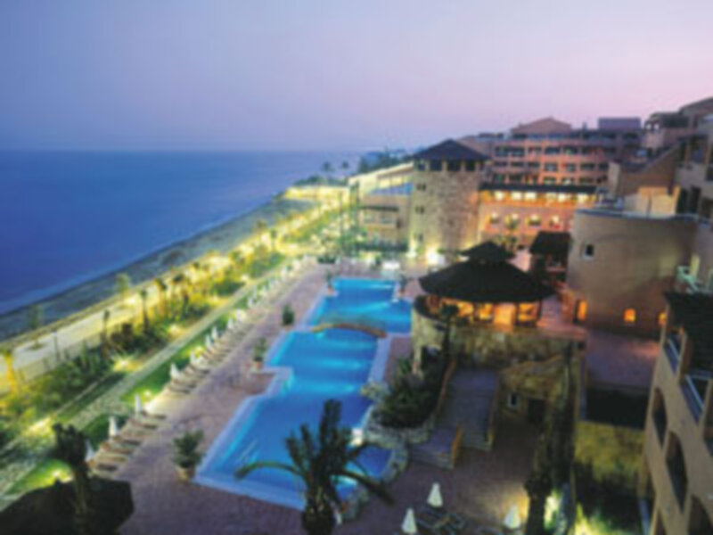 Imagen nocturna de un resort con piscinas al aire libre y edificios iluminados, frente al mar.
